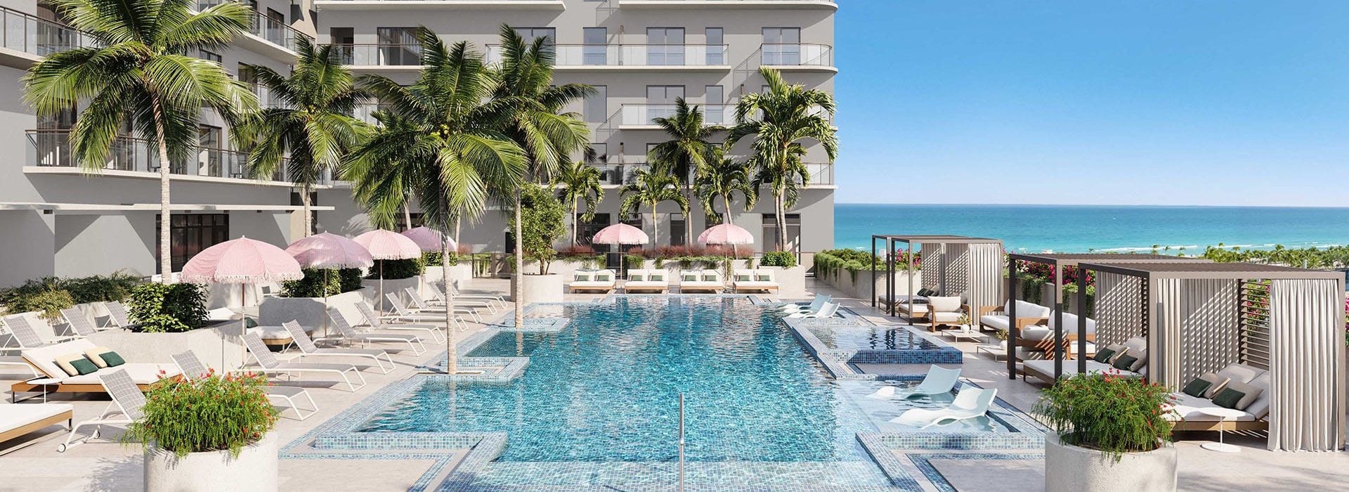 Pool area at a seaside resort with palm trees, pink umbrellas, and white loungers, facing the ocean.